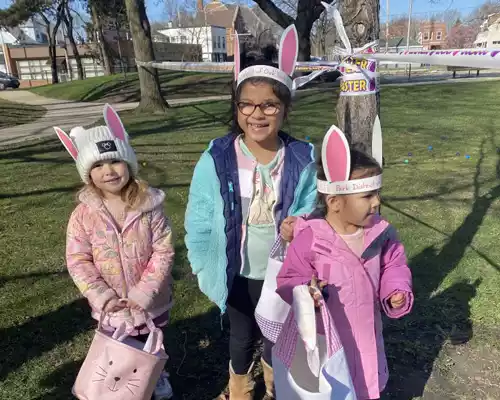 Three young girls wearing bunny ears and Easter-themed outfits, holding baskets, ready for an Easter egg hunt.