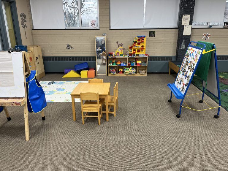 A children's play area with a small table and chairs, toy shelves, an easel, and soft building blocks.