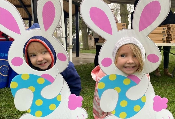 Two children pose with their faces through bunny-shaped cutouts holding colorful Easter eggs at the Egg Dash & Scramble