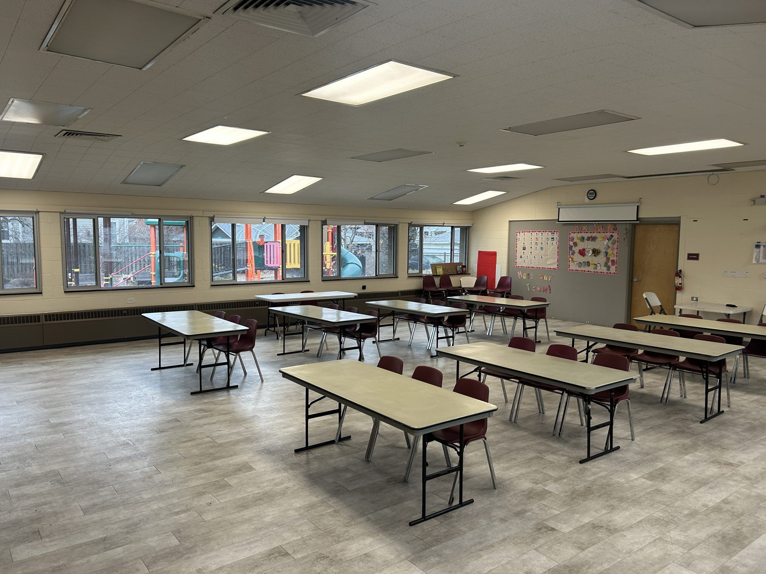 Tables and burgundy chairs facing the front of the room set up in a spacious room with wood doors and large windows.