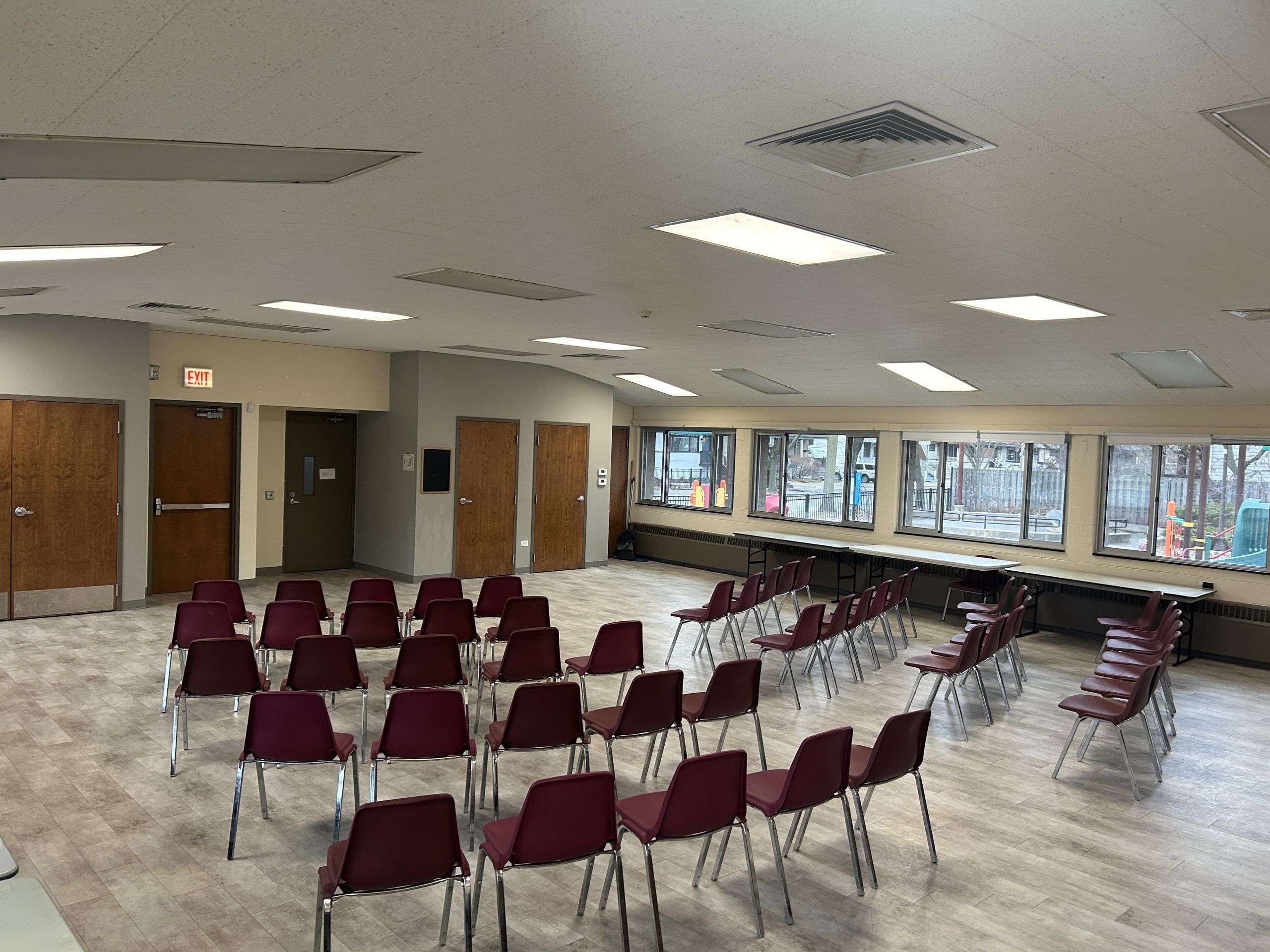 Rows of burgundy chairs facing the front of the room set up in a spacious room with wood doors and large windows.