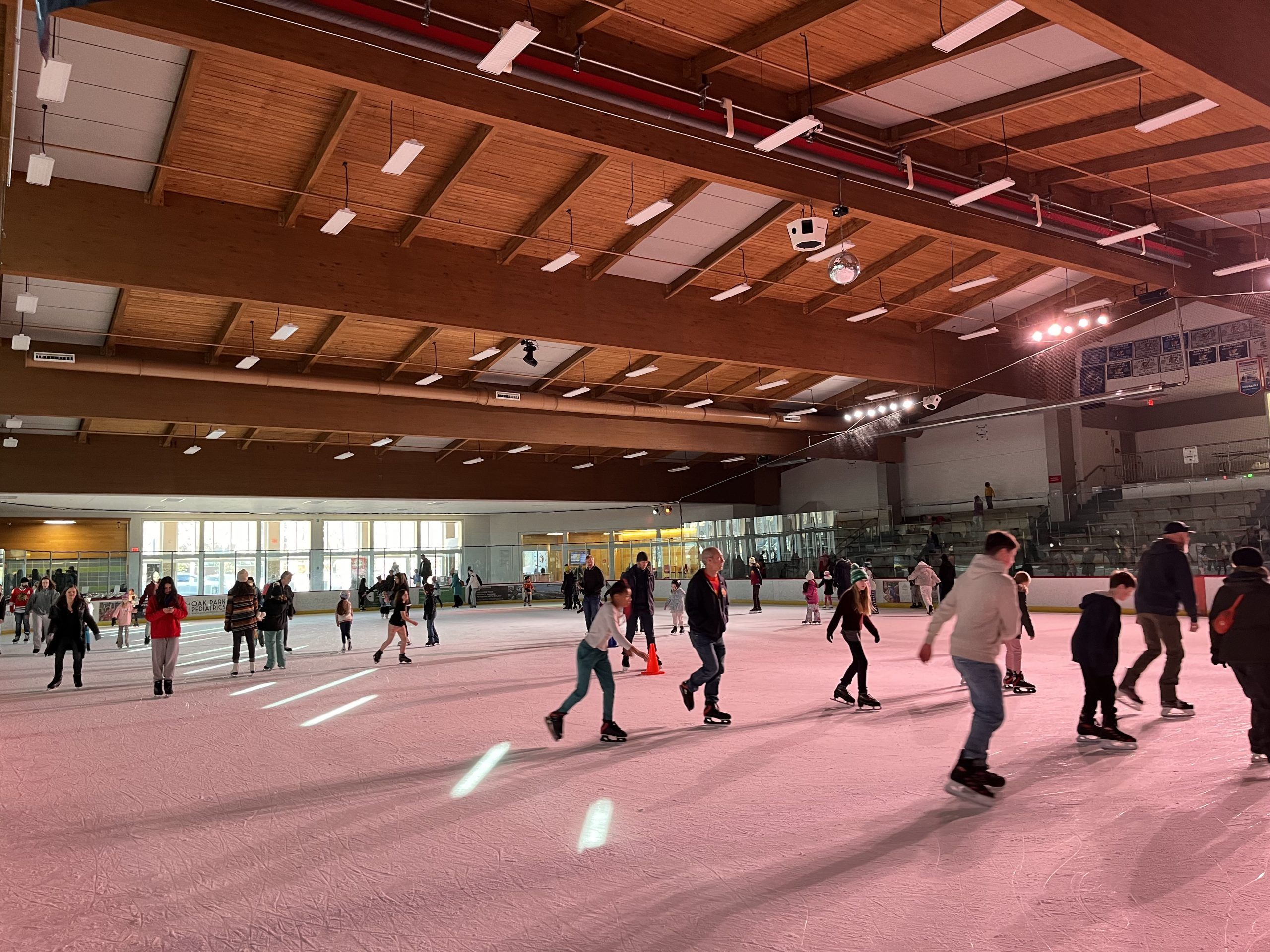 People ice skating indoors under a wooden beamed ceiling with bright lights and a disco ball.