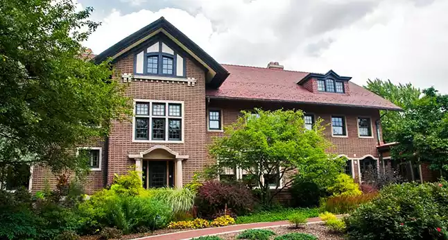 A large brick house with a red roof and many windows, surrounded by lush green trees and landscaping.