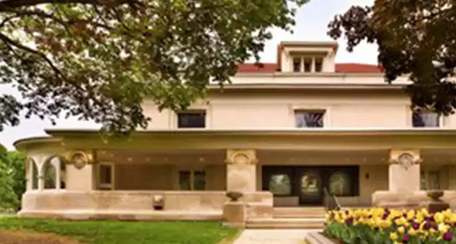 A large, light-colored house with a wide porch and red roof, surrounded by trees and flowers.