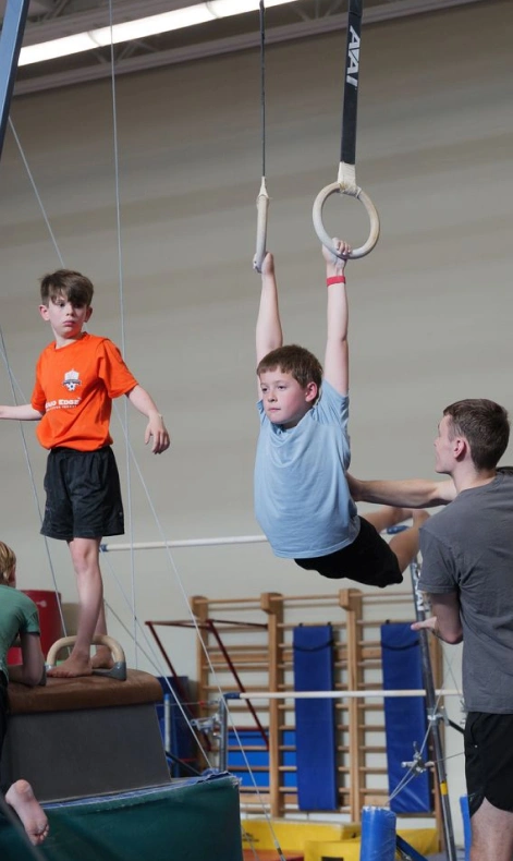 A boy practices gymnastics on rings with a coach spotting him. Another boy stands nearby.
