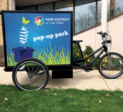 A three-wheeled bicycle with a large cargo box displaying "PARK DISTRICT of OAK PARK pop-up park" graphics.