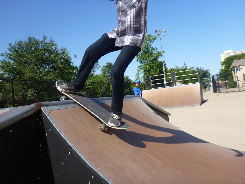 Skateboarder on a ramp at a mini skate park with a child in a blue helmet watching.