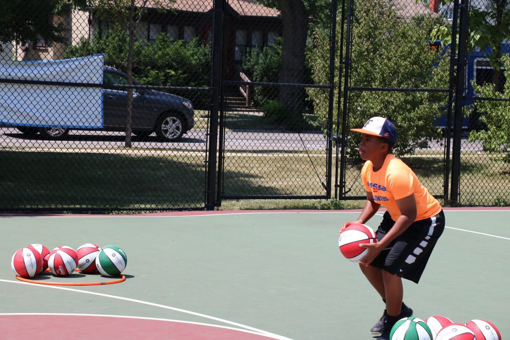 A young person in an orange shirt and baseball cap holds a basketball, ready to play on an outdoor court. Several other