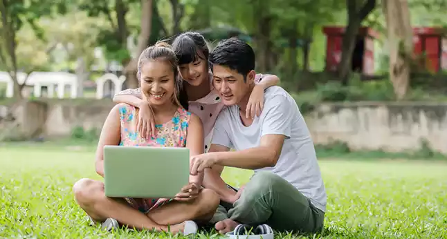 A family of three sits on grass in a park, looking at a laptop together. The mother and father are smiling, and the daughter