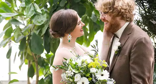 A smiling bride and groom embrace, holding a bouquet of white and green flowers, surrounded by lush greenery at the Oak Park