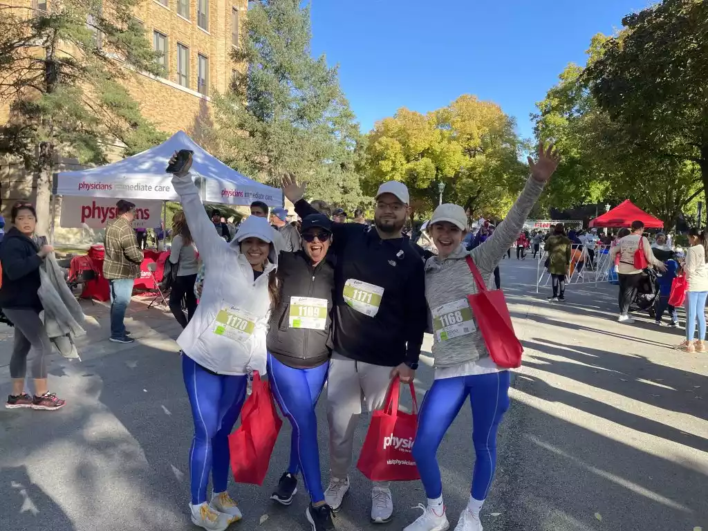 Four participants in a race smile and wave, wearing bibs 1170, 1169, 1171, and 1168. A Physicians Immediate Care tent is
