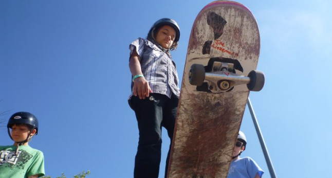 Three young skateboarders in helmets stand against a bright blue sky, one holding a worn skateboard.